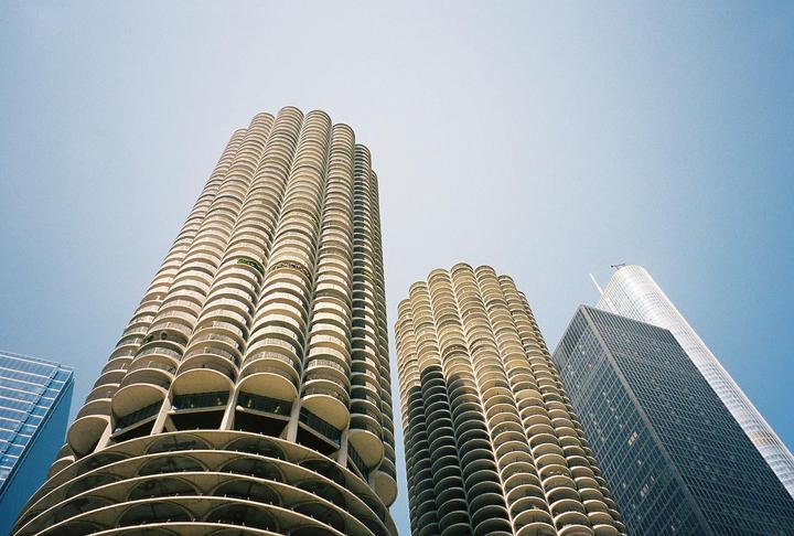 Two cylindrical high-rise buildings with many balconies stand next to other skyscrapers under a clear sky.