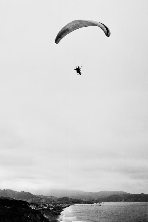 A paraglider is visible in the sky above a coastline with mountains in the distance.