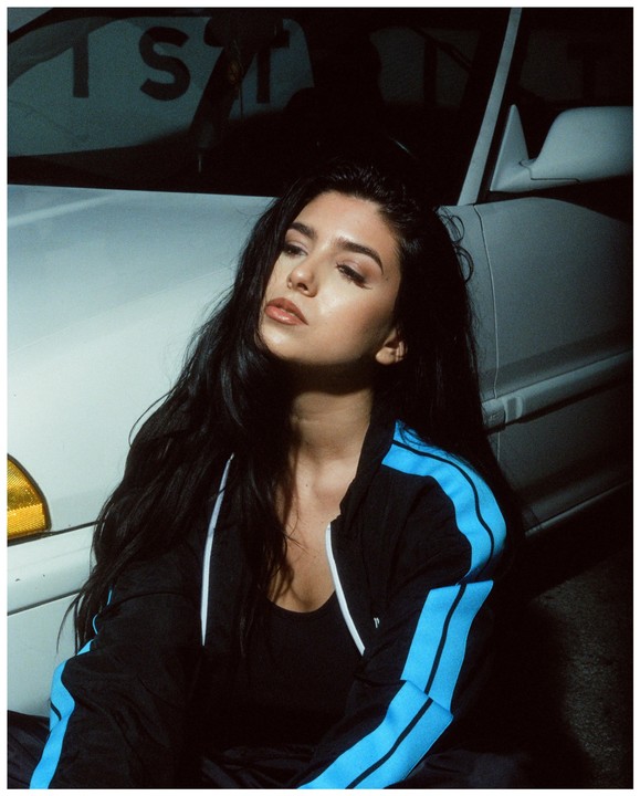 A woman with long dark hair sits in front of a white car at night.