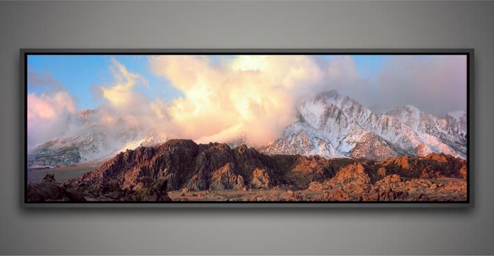 Clouds hang over snow-capped mountains and rocky hills illuminated by the golden hour light.