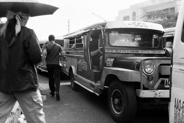 A man walks past a jeepney on a street while another person holds an umbrella in the foreground.