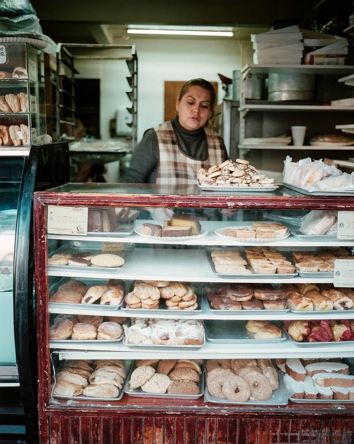 A woman stands behind a glass display case filled with pastries in a pastry shop.