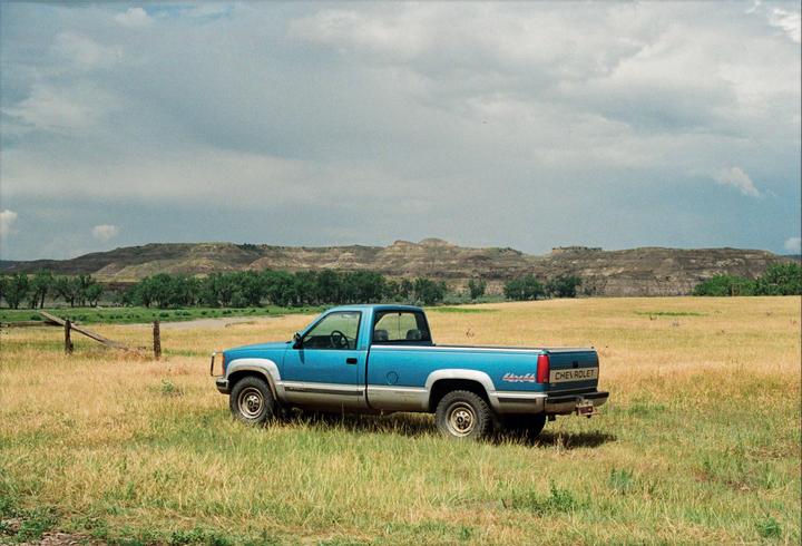 A blue Chevrolet pickup truck is parked in a grassy field with hills and trees in the background under a cloudy sky.