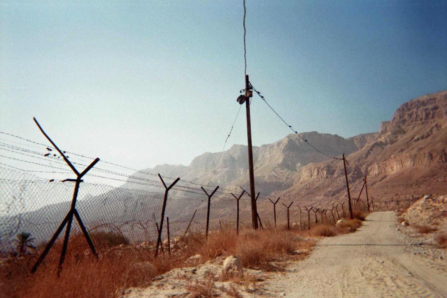 A dirt road runs alongside a barbed wire fence with mountains in the background under a clear sky.