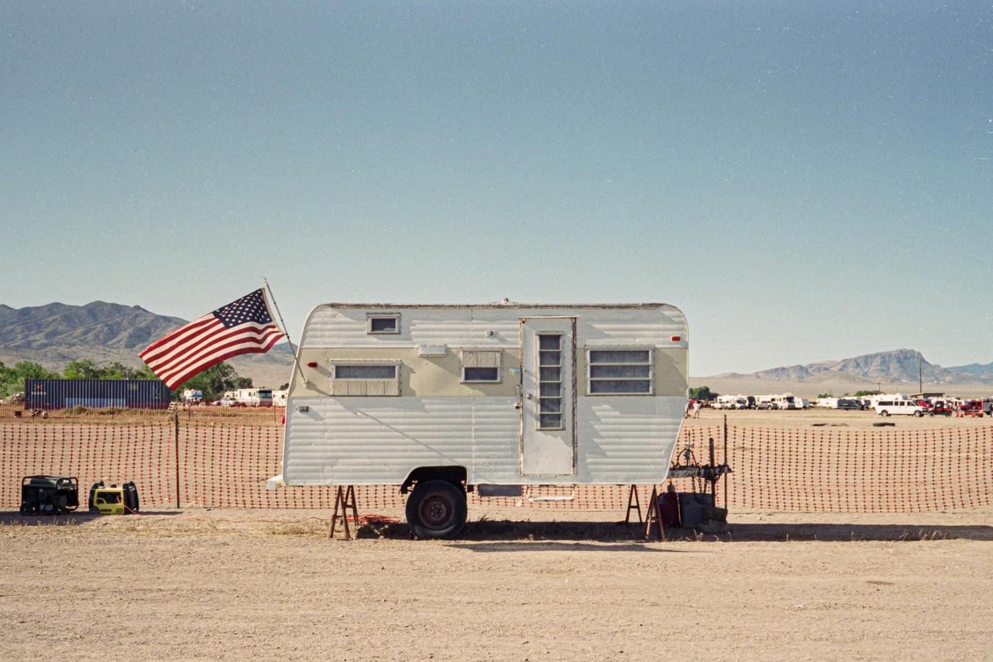 An American flag flies next to a white travel trailer in a desert landscape with mountains in the distance.