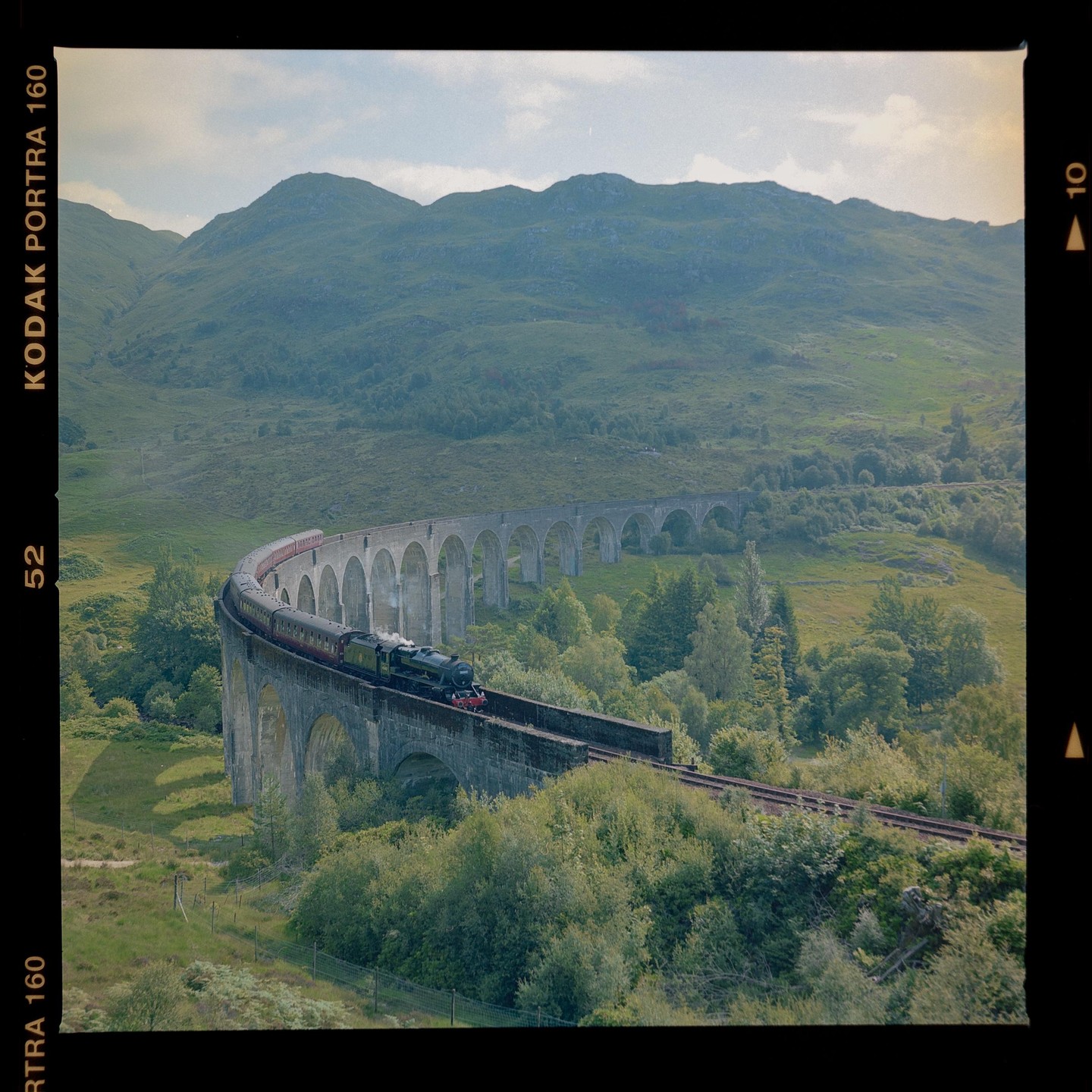 A steam train crosses a long, arched viaduct through a green, mountainous landscape.