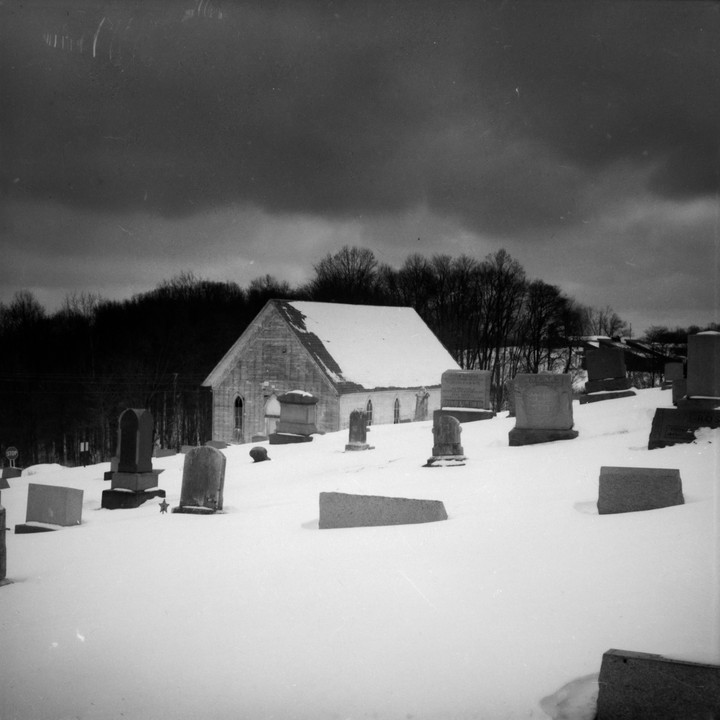 A snow-covered church stands in a cemetery under a dark, cloudy sky.