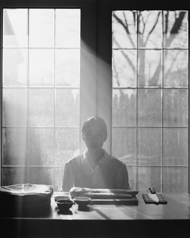 A woman sits at a table with a piece of salmon, bowls, and a knife, with light streaming through a window behind her.