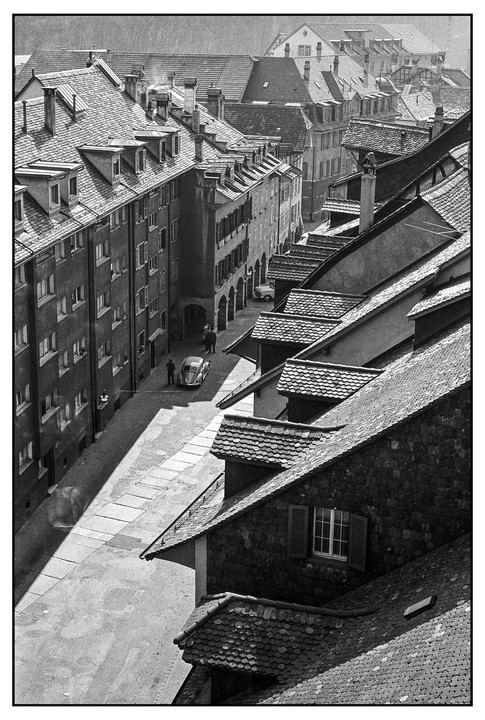 A high-angle view shows a street with a car and people, surrounded by buildings with many roofs.