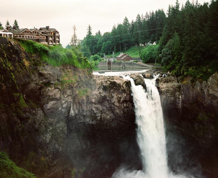 A large waterfall cascades into a rocky gorge with a building on the cliff edge and a forest in the background.