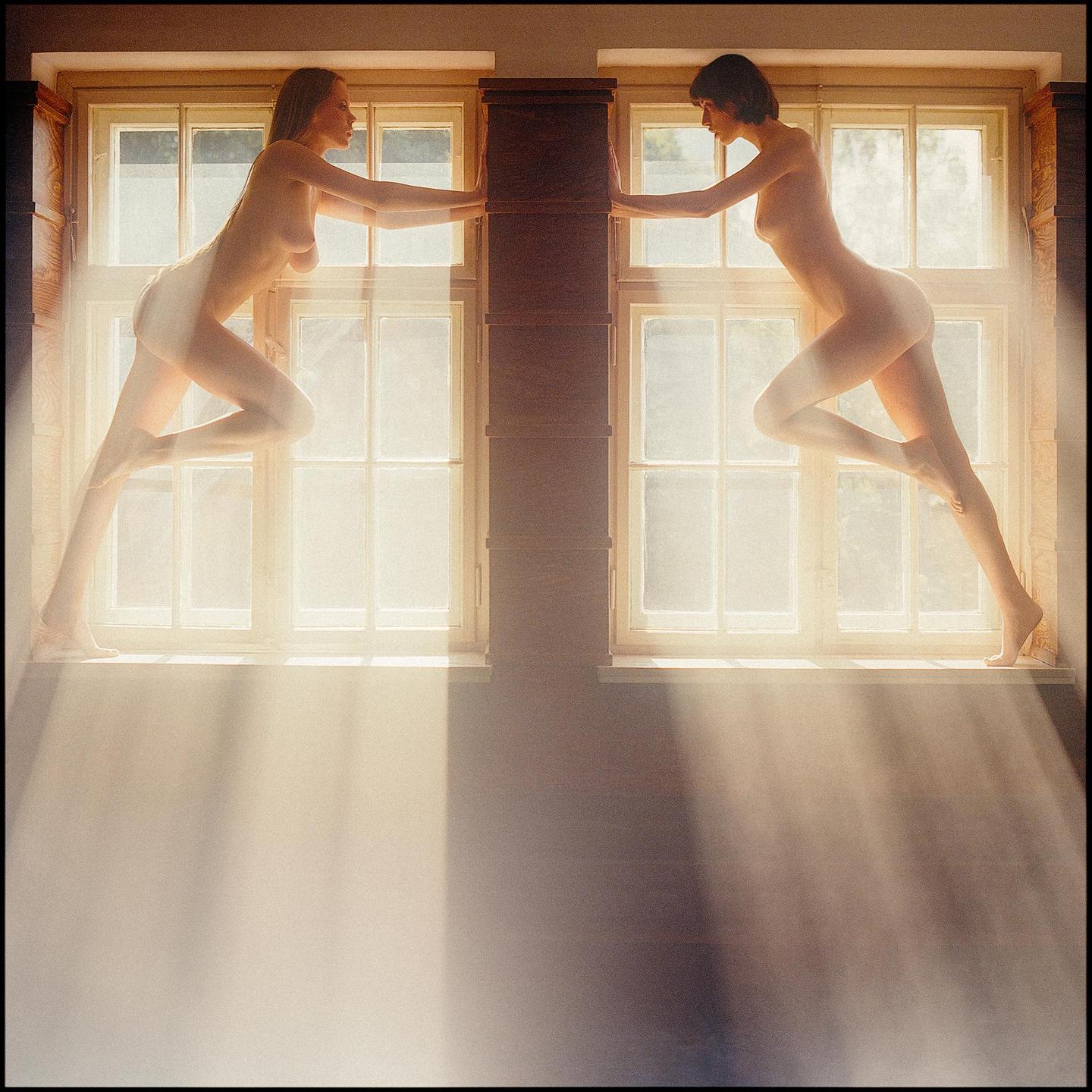 Two women pose nude on window sills, illuminated by rays of sunlight streaming through the windows.