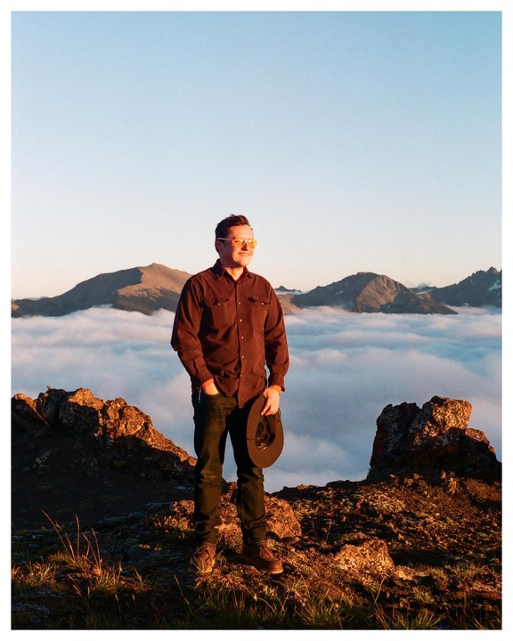 A man stands on a rocky mountain peak above a sea of clouds, holding a hat.