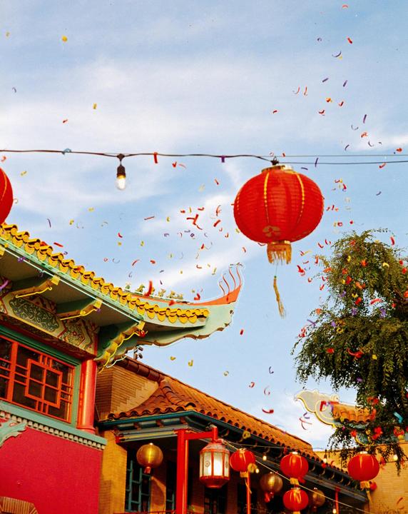 Confetti falls over a street in Chinatown with red lanterns and ornate rooftops under a blue sky.