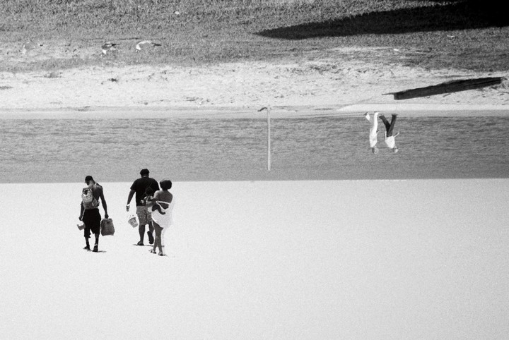 Three people walk away from the viewer on a bright beach toward the water, with two birds flying above the water and two people upside down in the distance.