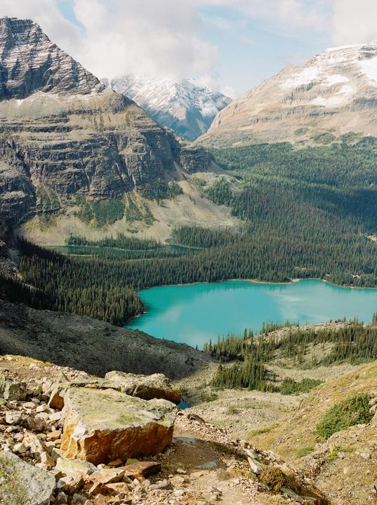 A turquoise lake is nestled among a dense forest and rocky mountains with snow-capped peaks.