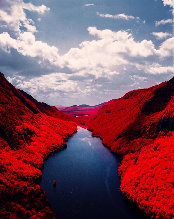 A river flows through a valley with red foliage on the mountainsides under a cloudy sky.