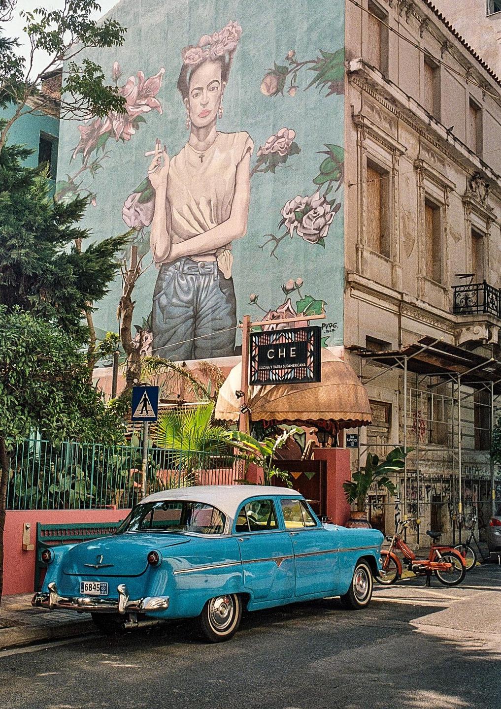 A blue vintage car is parked on a street next to a building with a large mural of a woman smoking.