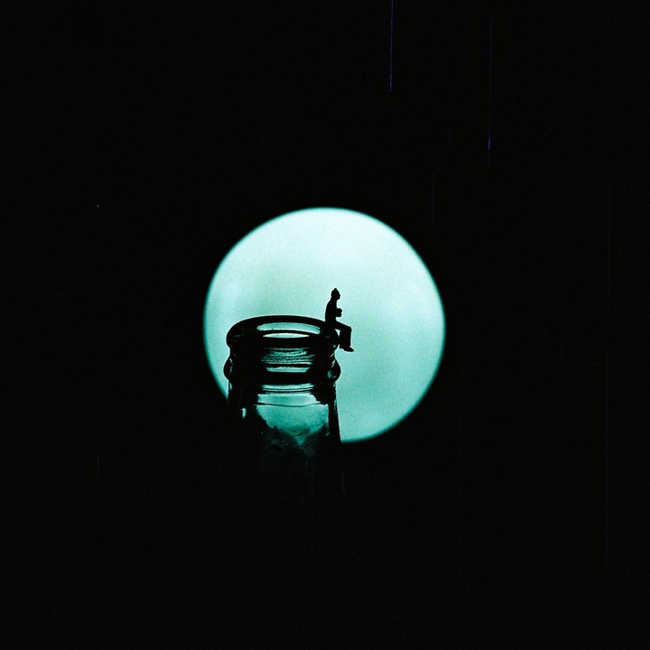 A small figure sits on the rim of a bottle with a bright circle in the background.