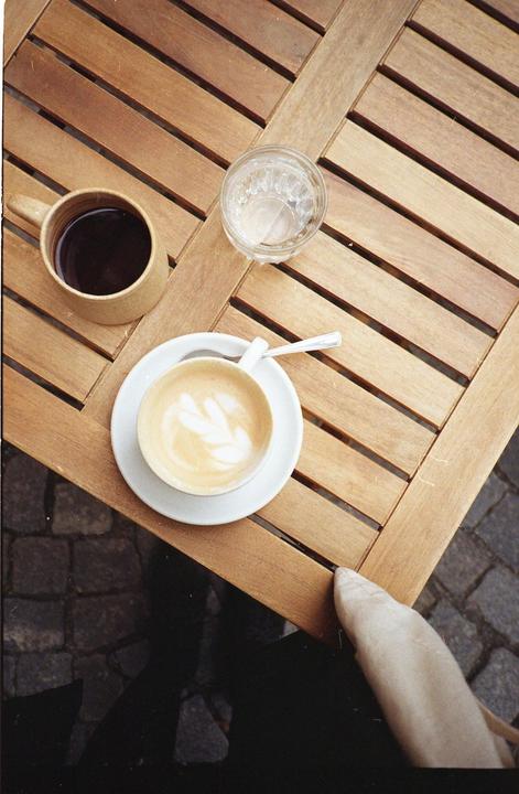 A cup of coffee, a latte, and a glass of water sit on a slatted wooden table next to a person's leg.