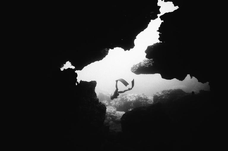 A woman freedives through a shallow cave, viewed from inside the cave looking out.