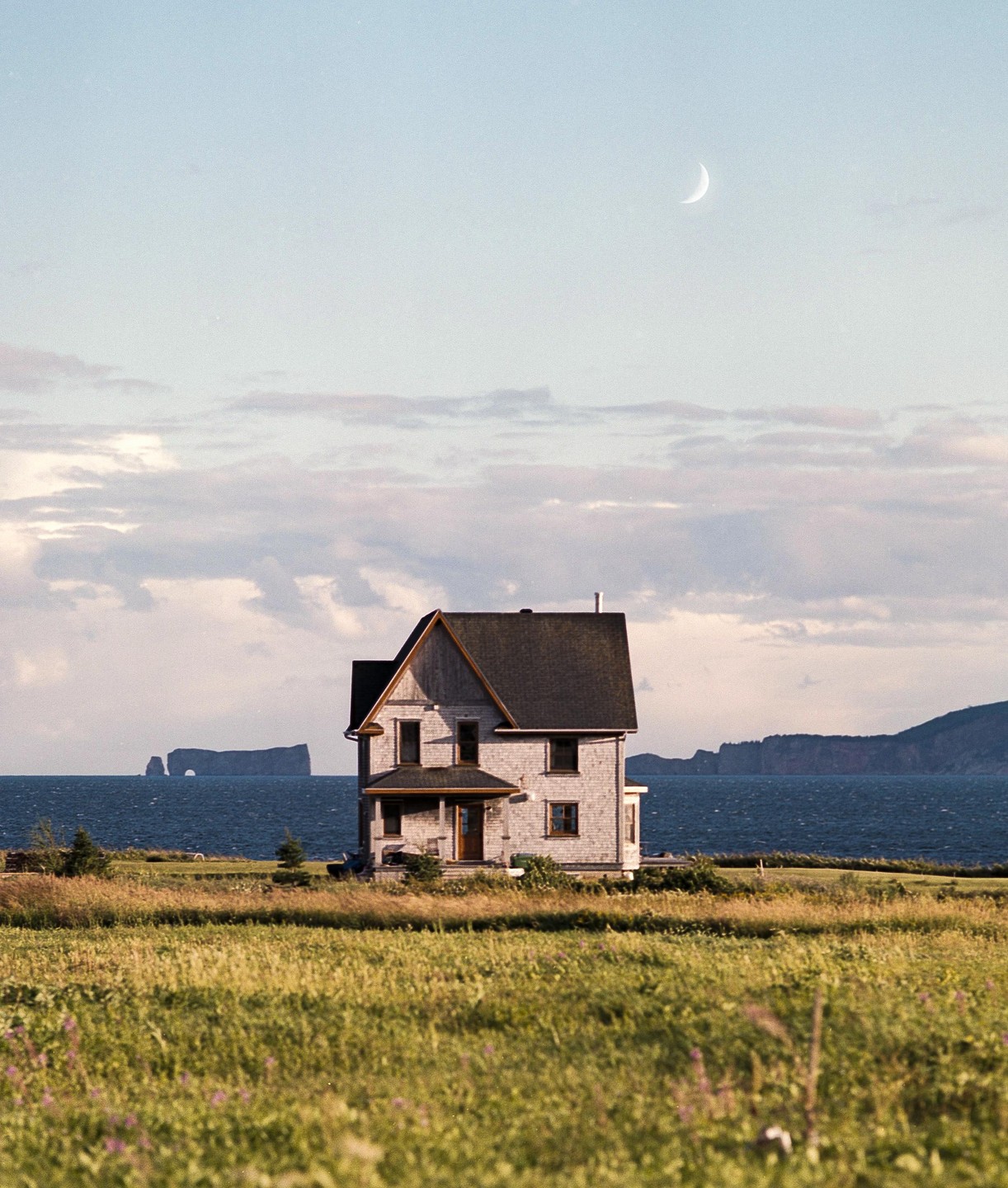 A house stands in a field overlooking the ocean with a crescent moon in the sky.