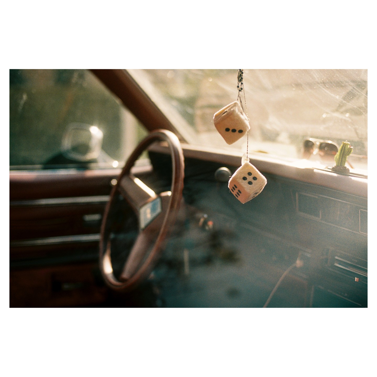 Two fuzzy dice hang from the rearview mirror of a car, with a steering wheel visible on the left.