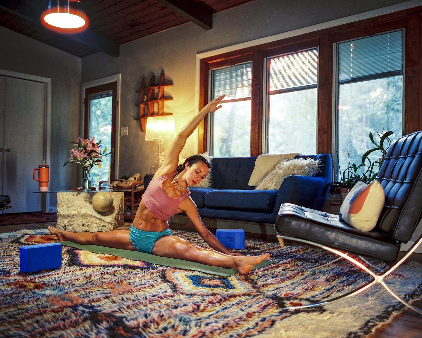 A woman stretches on a yoga mat in a living room with a colorful rug.