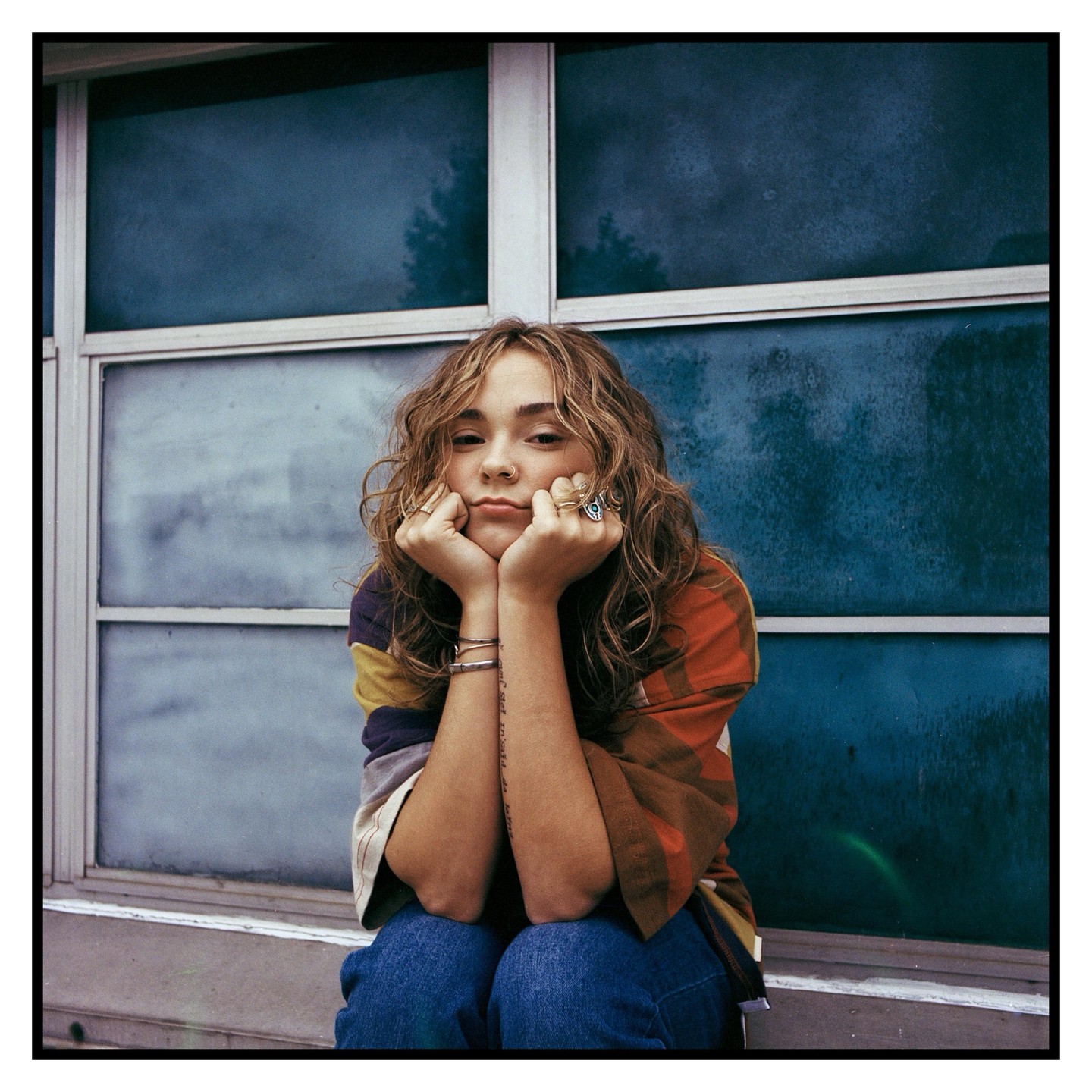 A young woman with curly hair and rings on her fingers rests her chin on her hands in front of a window.