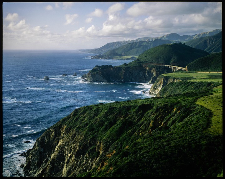 A bridge crosses a deep canyon along a rugged coastline with green hills and a blue ocean.