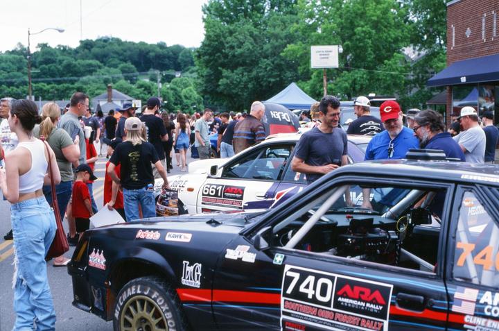 A crowd of people gathers around two rally cars on a street lined with trees and buildings.