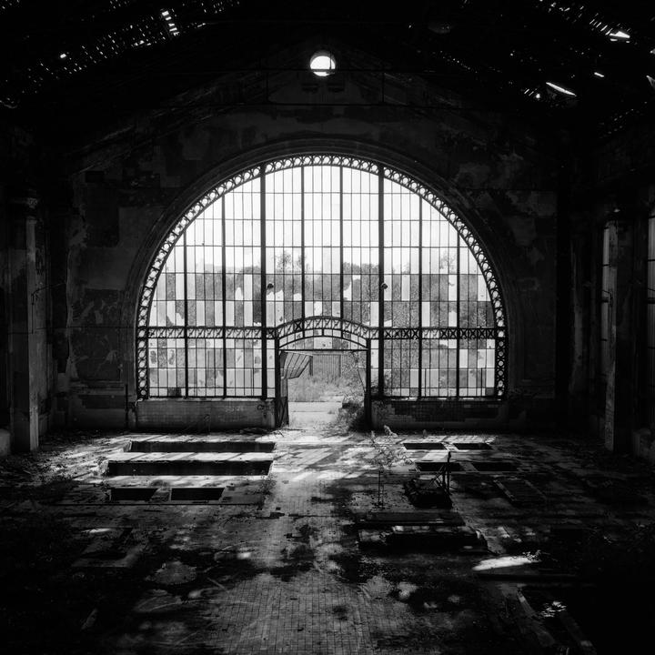 Sunlight streams through a large arched window into a dark, abandoned industrial building.