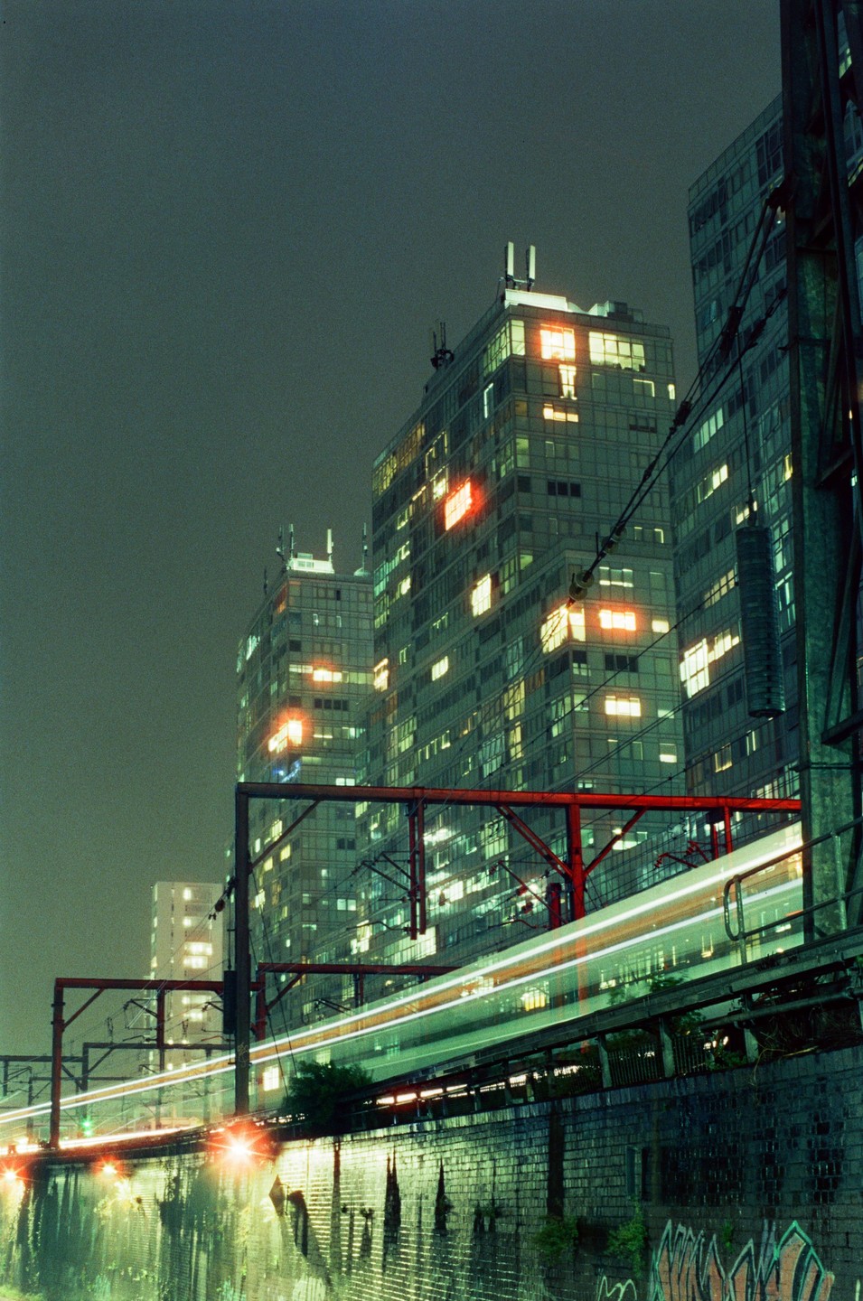 A light trail from a train passes by tall buildings at night.