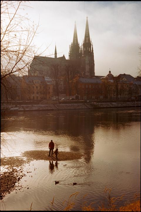 A man and a child stand on a small island in a river with a city and cathedral in the background.