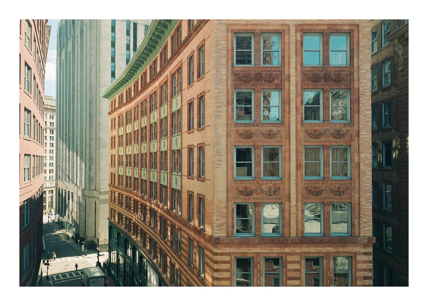A curved brick building with many windows stands next to a tall, light-colored building, with a street and other buildings visible below.