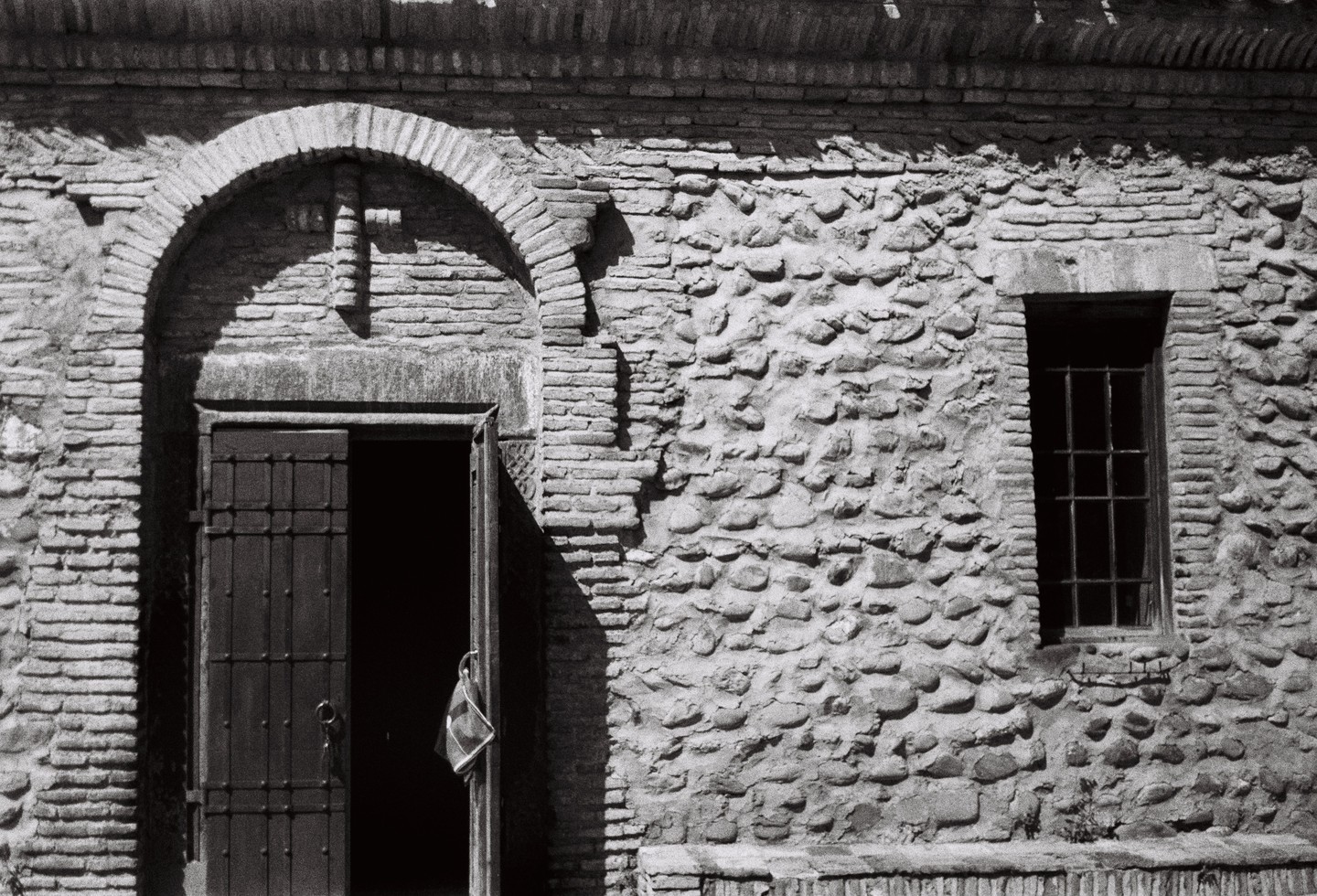 An open door with a small bag hanging from it is next to a barred window on a stone and brick wall.