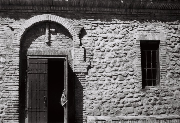 An open door with a small bag hanging from it is next to a barred window on a stone and brick wall.