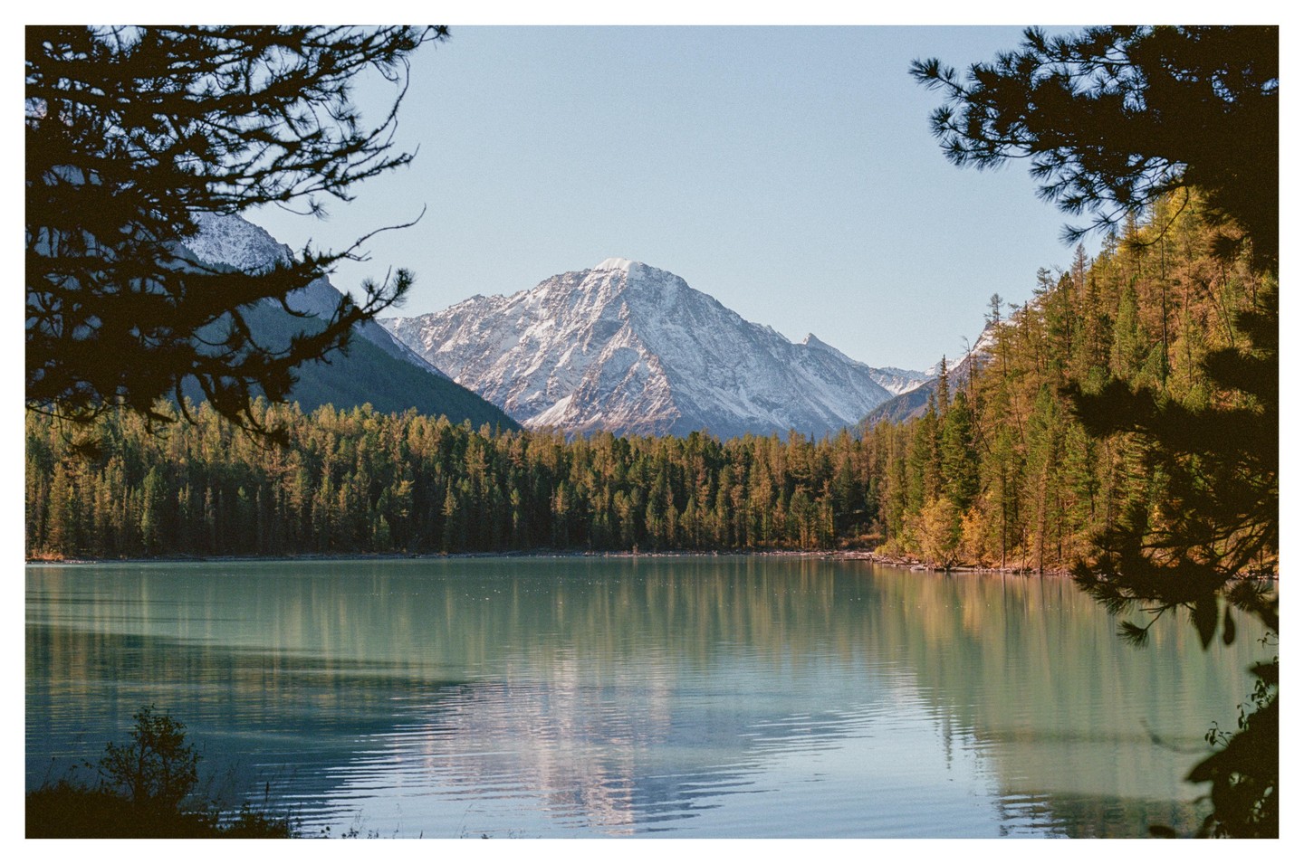 A lake is surrounded by a forest with a snow-capped mountain in the background under a clear sky.