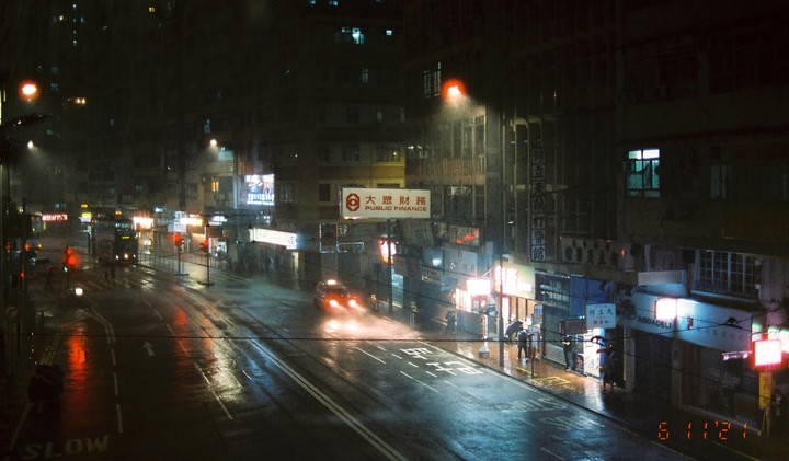 A street is wet from rain at night, with cars and people visible under glowing signs and streetlights.