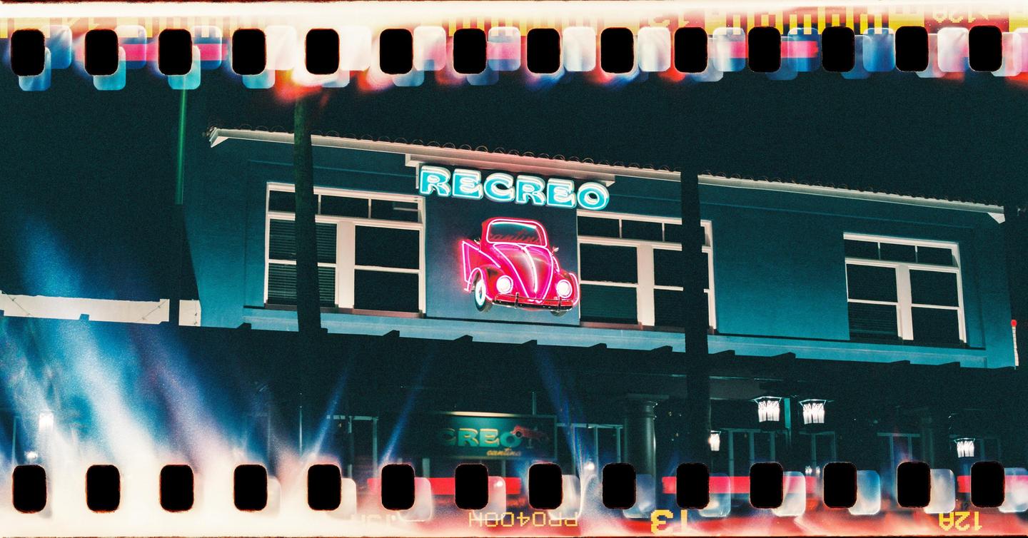 A neon sign of a car and the word "RECREO" is displayed on the side of a building at night.