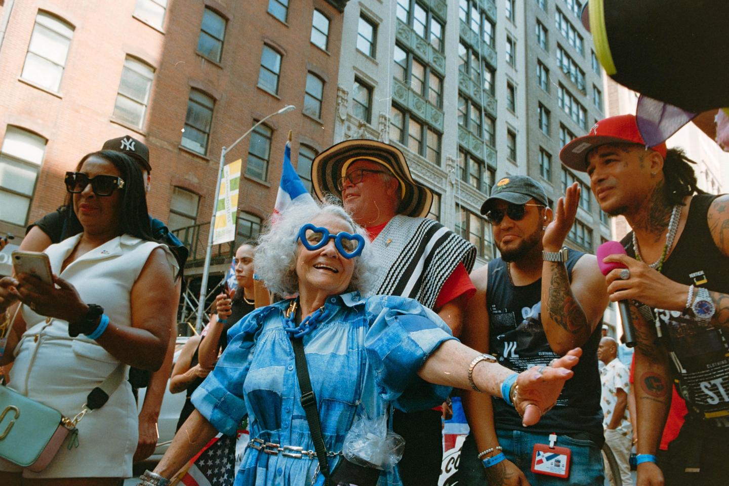 A woman in heart-shaped glasses and a blue dress smiles while standing in a crowd on a city street.