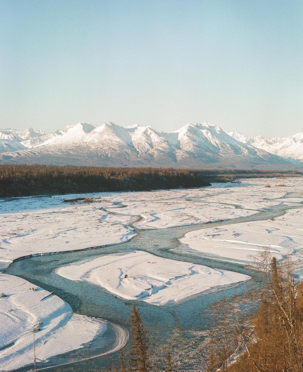 A wide river with snow-covered banks flows through a valley with a forest and snow-capped mountains in the background.
