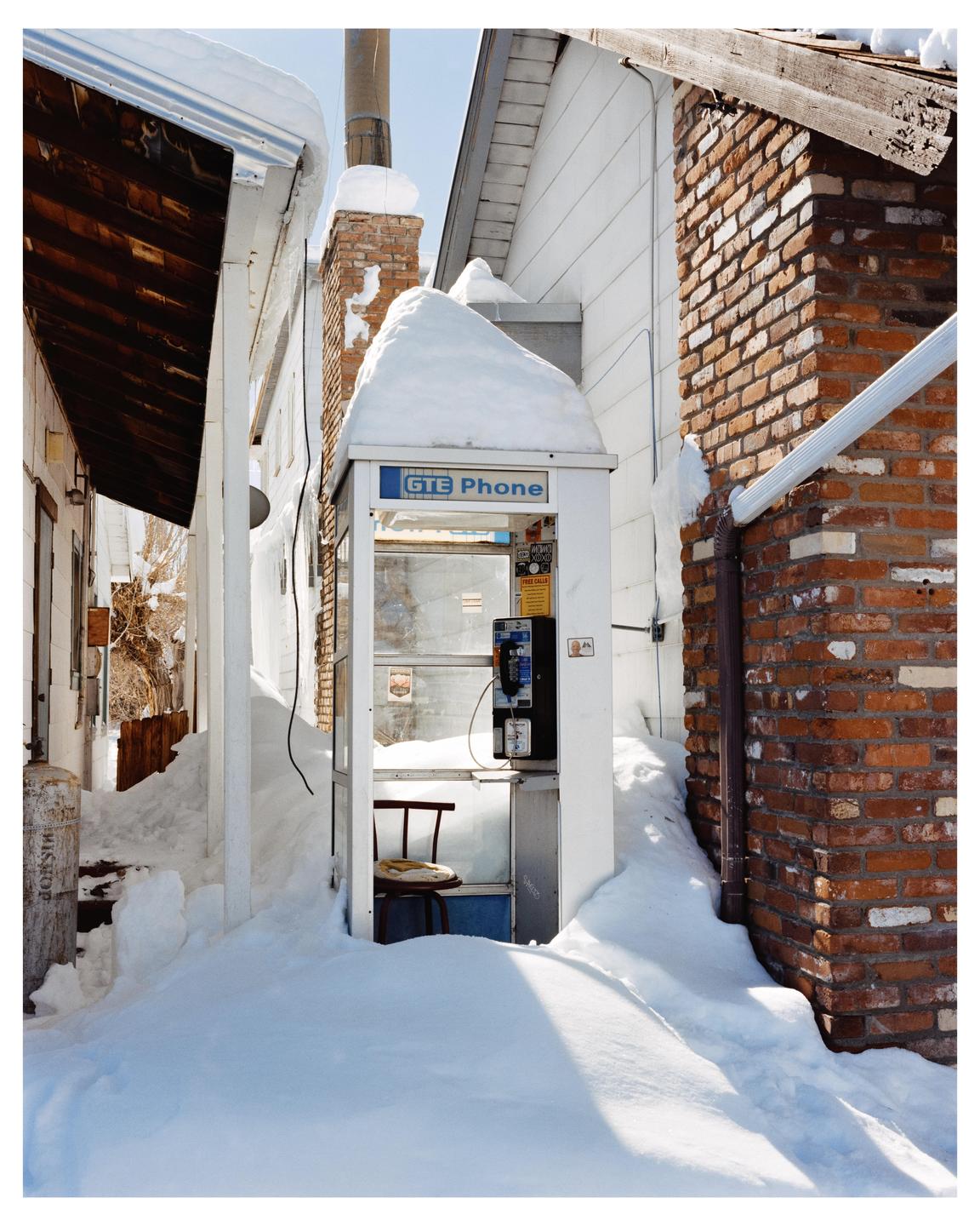 A white telephone booth is surrounded by deep snow between two buildings.