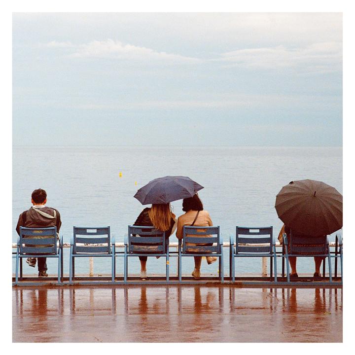 Four people sit on blue chairs facing the ocean, two of them holding umbrellas.