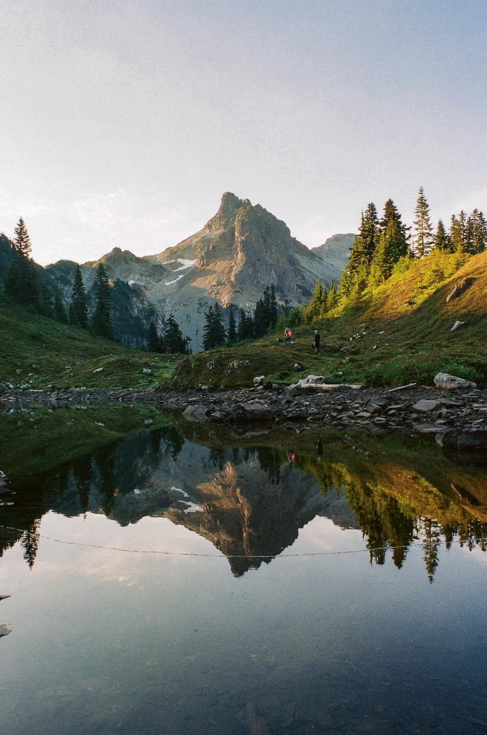 A mountain peak is reflected in a still lake with two people walking on a grassy hill.