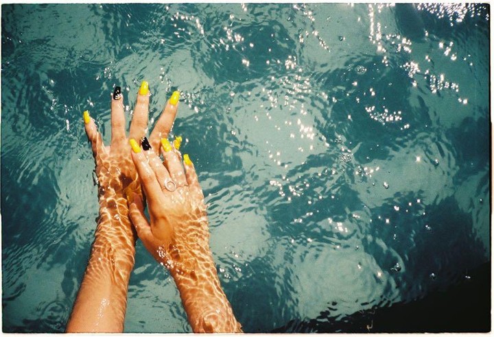 A person's hands with yellow and black nail polish are submerged in sparkling blue water.
