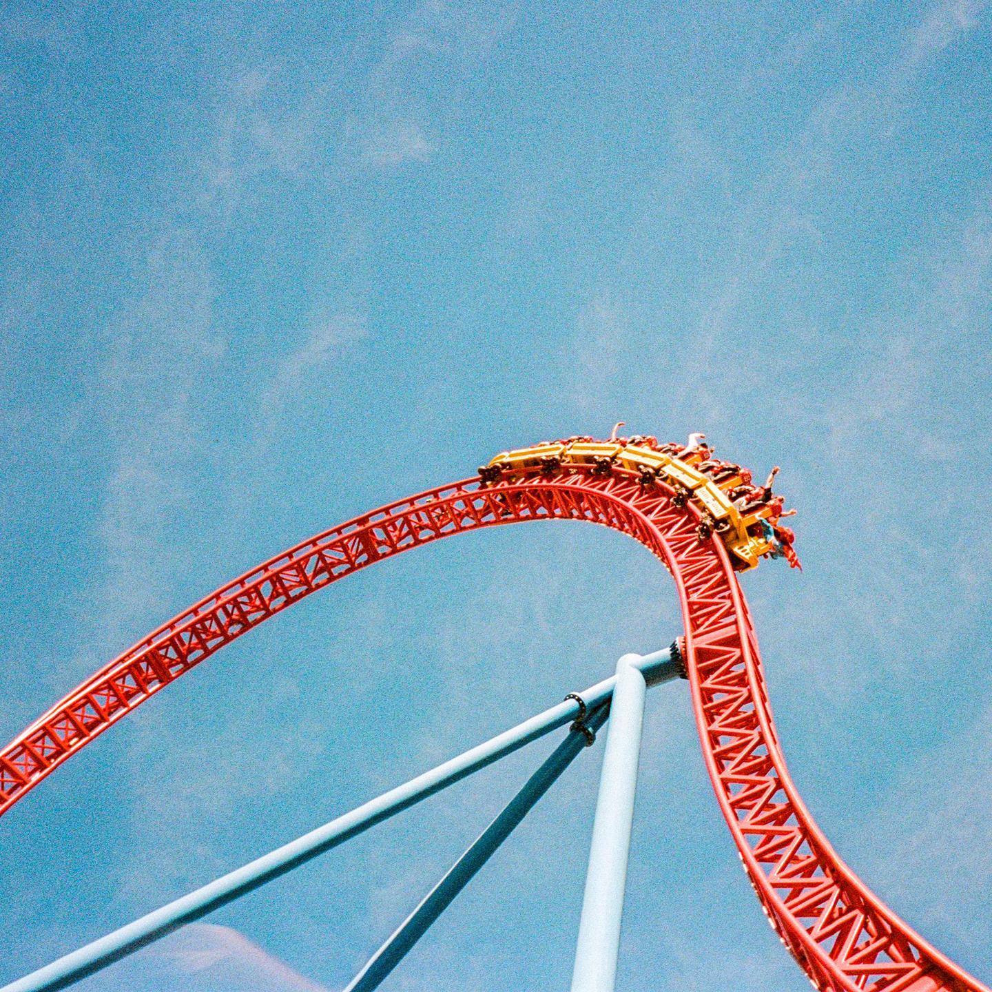 A yellow roller coaster car full of people is at the top of a red track against a blue sky.