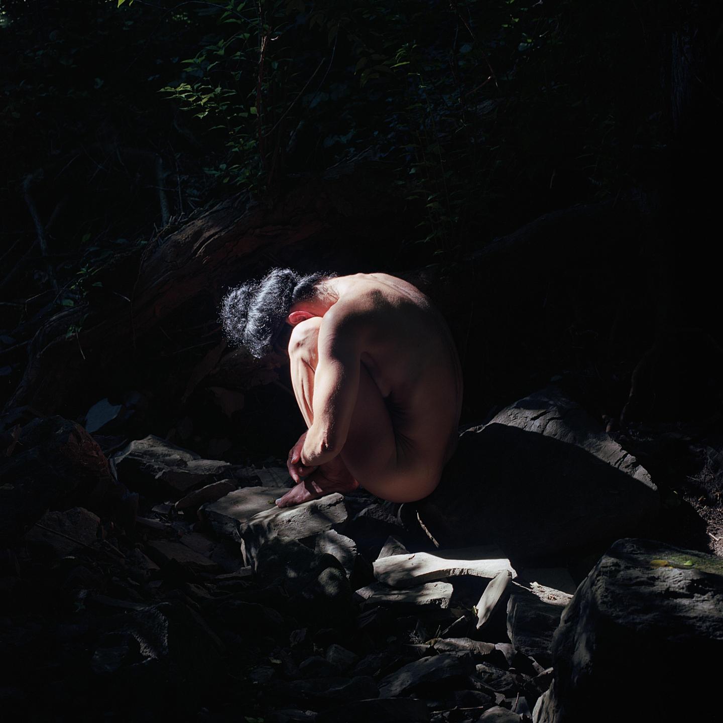 A nude woman sits curled up on a rock in a dark forest, illuminated by a shaft of light.