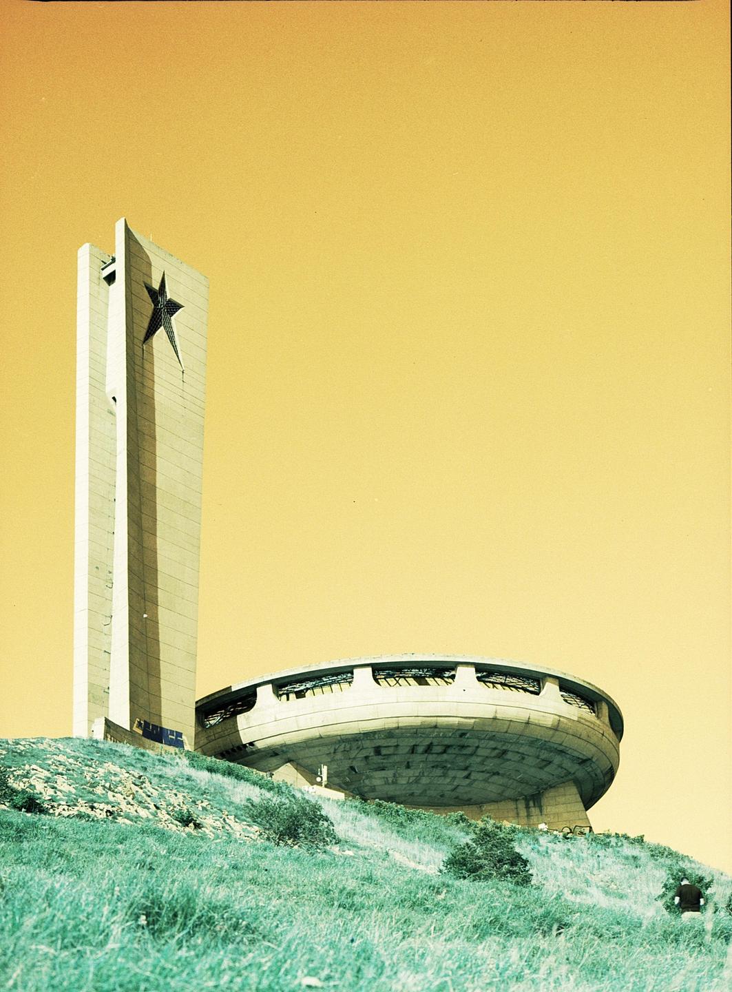 A concrete tower with a star and a circular concrete building are on a grassy hill under a yellow sky.