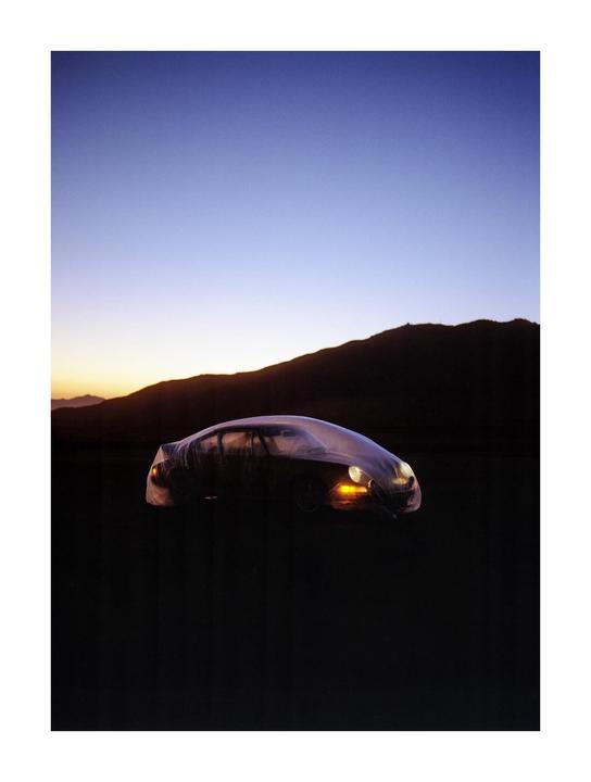 A car covered in a translucent sheet is parked in a dark landscape with mountains silhouetted against a colorful sky.