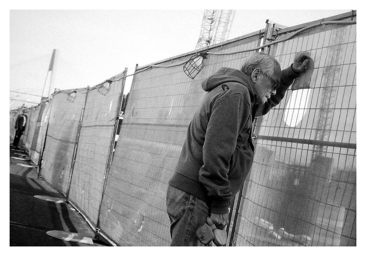 A man in a hoodie leans against a chain-link fence, looking down, with another man in the distance.